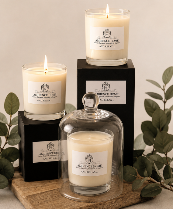 Three Ambience Home candles in glass vessels with white labels are arranged on a wooden tray with black boxes and green leaves against a warm neutral background.