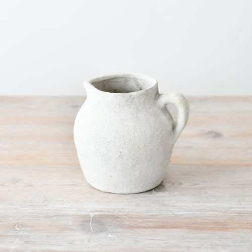 Textured white ceramic jug vase with a rounded shape and single handle, featuring a rustic stone-effect finish on a wooden surface.