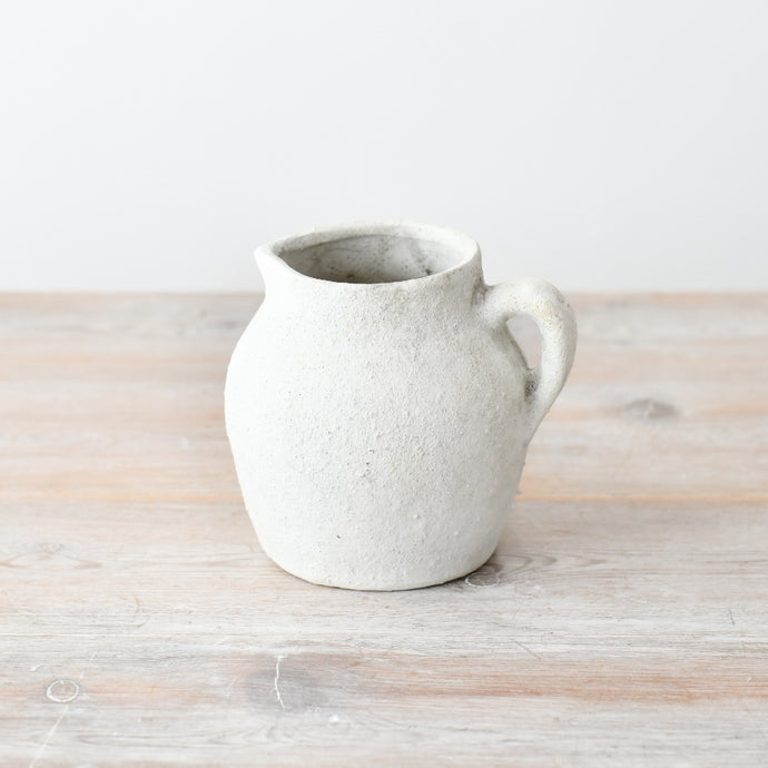 Textured white ceramic jug vase with a rounded shape and single handle, featuring a rustic stone-effect finish on a wooden surface.
